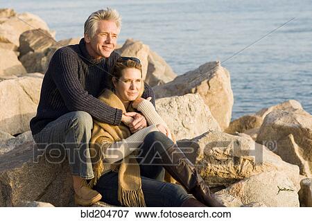 Stock Photo - Caucasian couple sitting on rocky beach. Fotosearch