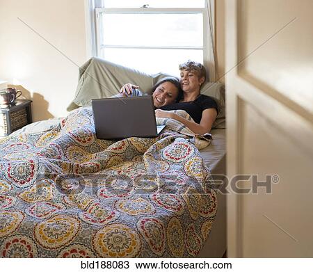 Stock Image - Caucasian lesbian couple watching laptop in bed. Fotosearch
