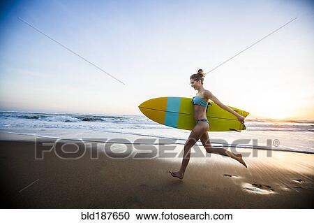 Stock Image - Caucasian woman carrying surfboard on beach. Fotosearch