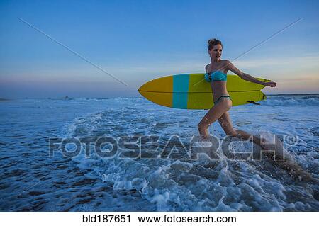 Stock Image - Caucasian woman carrying surfboard on beach. Fotosearch