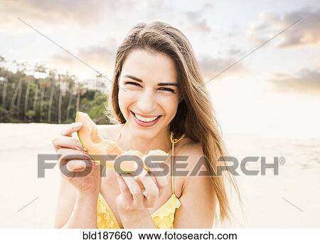 Stock Image - Caucasian woman eating fruit on beach. Fotosearch