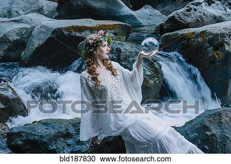 Stock Image - Caucasian woman holding crystal ball at river waterfall. Fotosearch