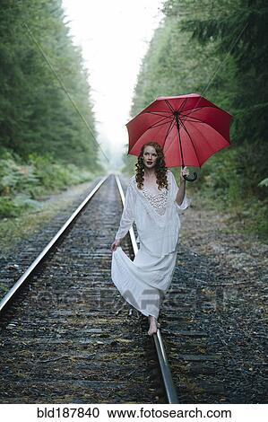 Stock Image - Caucasian woman walking on train tracks with umbrella in forest. Fotosearch