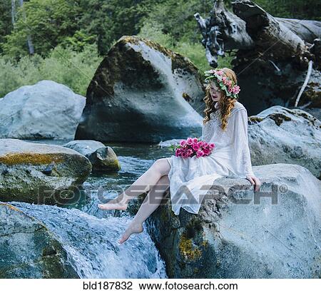 Stock Image - Caucasian woman wearing flower crown on rock at river waterfall. Fotosearch