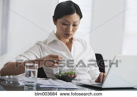 Picture - Chinese businesswoman eating salad at desk. Fotosearch