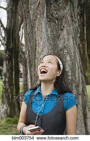 Picture - Chinese girl listening to headphones against tree. Fotosearch