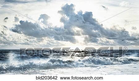 Stock Image - Cloudy sky over stormy waves on beach. Fotosearch