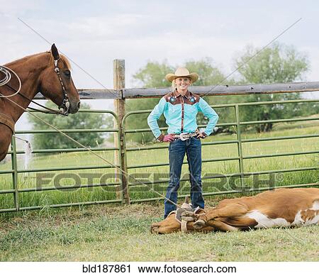 Stock Image - Cowgirl smiling with tied cattle on ranch. Fotosearch