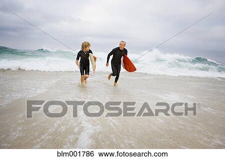 Stock Photograph - Father and son running out of ocean with surfboards. Fotosearch