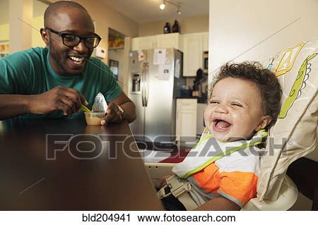 Stock Image - Father feeding baby at table. Fotosearch