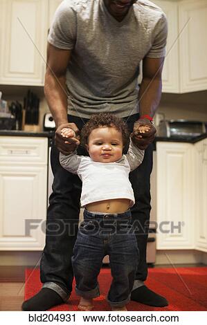Stock Image - Father helping baby walk in kitchen. Fotosearch