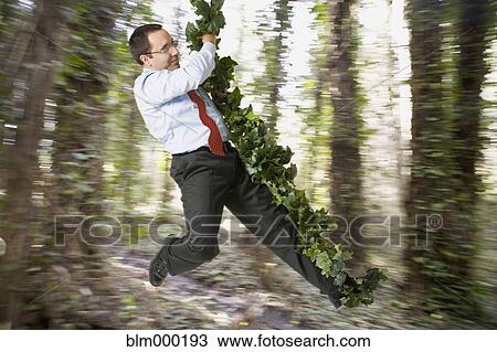 Stock Image - Hispanic businessman swinging on forest vine. Fotosearch