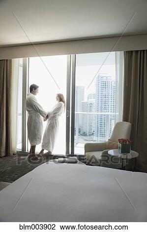Stock Image - Hispanic couple in bathrobes holding hands by window. Fotosearch