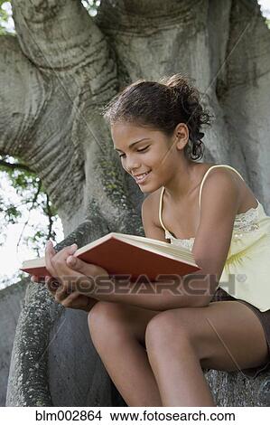 Picture - Hispanic girl reading book on tree trunk. Fotosearch