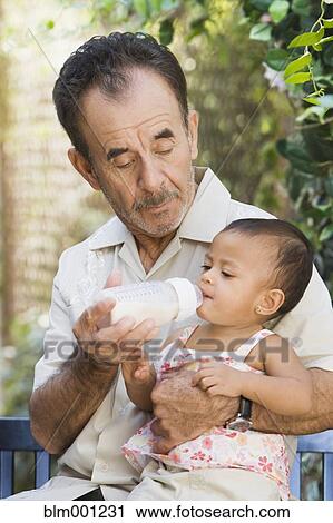 Stock Image - Hispanic grandfather feeding bottle to granddaughter. Fotosearch