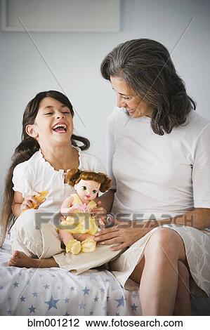 Stock Image - Hispanic grandmother and granddaughter with doll in bedroom. Fotosearch
