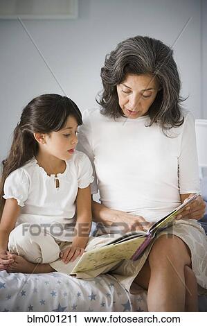 Stock Image - Hispanic grandmother reading to granddaughter in bedroom. Fotosearch
