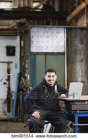 Picture - Hispanic male metalworker in shop with laptop. Fotosearch