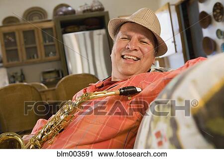 Stock Image - Hispanic man holding saxophone on sofa. Fotosearch