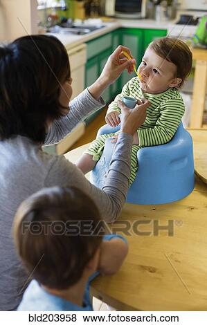Stock Photo - Hispanic mother feeding toddler in high chair. Fotosearch