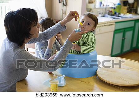 Stock Photo - Hispanic mother feeding toddler in high chair. Fotosearch