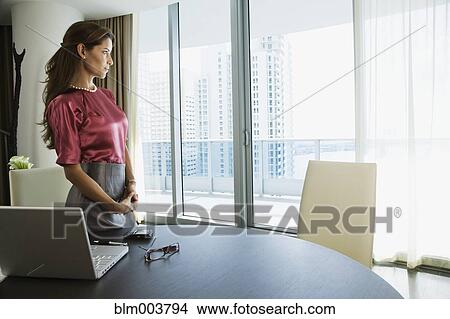 Picture - Hispanic woman standing next to laptop and looking out window. Fotosearch