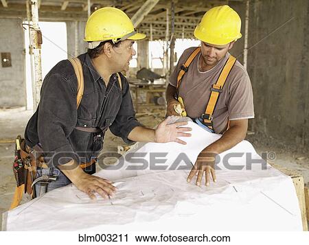Stock Image - Hispanic workers looking at blueprints on construction site. Fotosearch