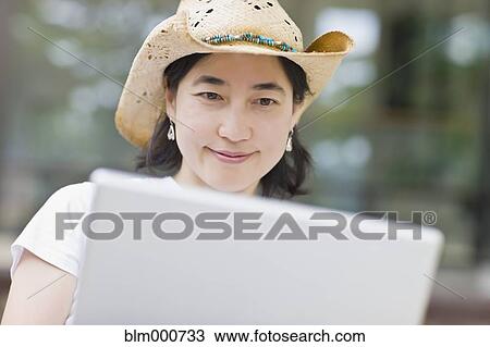 Stock Image - Japanese woman typing on laptop wearing cowboy hat. Fotosearch