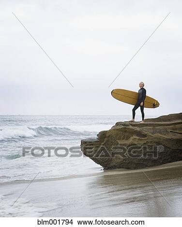 Man holding surfboard on rocks at beach View Large Photo Image Picture - Man holding surfboard on rocks at beach. Fotosearch