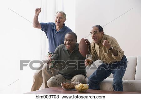 Stock Image - Men watching football game in living room with snacks. Fotosearch