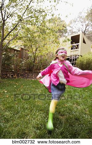 Stock Image - Middle Eastern girl wearing cape and mask running in backyard. Fotosearch