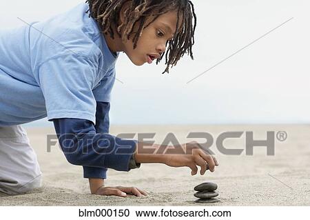 Stock Image - Mixed Race boy stacking stones on sand. Fotosearch