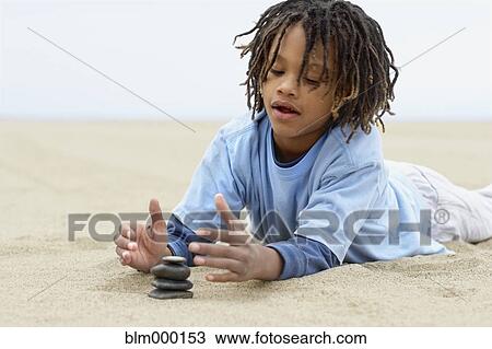 Stock Image - Mixed Race boy stacking stones on sand. Fotosearch