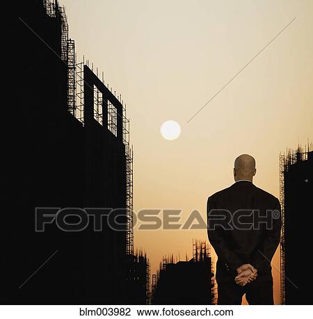 Stock Image - Mixed race businessman looking at sun setting in sky. Fotosearch