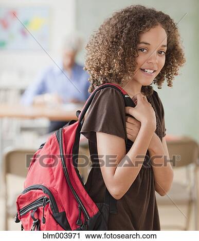 Stock Image - Mixed race girl holding backpack in classroom. Fotosearch