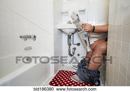 Stock Image - Mixed race man reading newspaper on toilet. Fotosearch