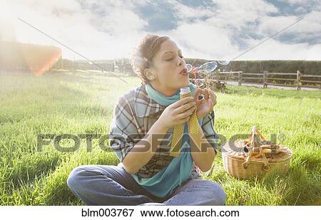 Stock Photo - Mixed race woman blowing bubbles in sunny field. Fotosearch