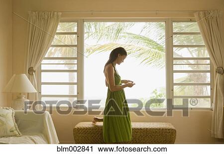 Stock Image - Mixed race woman looking at cell phone near window. Fotosearch