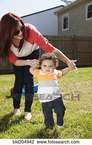 Stock Image - Mother helping baby walk in backyard. Fotosearch