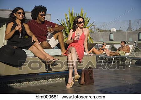 Stock Image - Multi-ethnic friends drinking cocktails poolside. Fotosearch