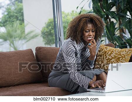 Stock Image - Nervous African woman typing on laptop in living room. Fotosearch