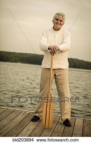 Picture - Older Caucasian man holding oar on wooden dock. Fotosearch