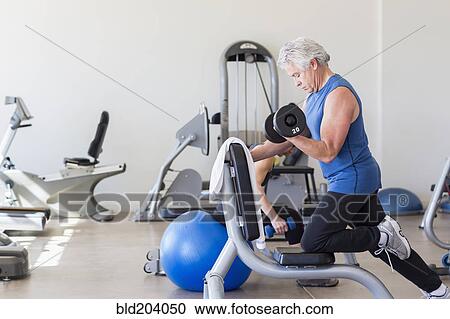 Stock Image - Older Hispanic man lifting weights in gym. Fotosearch