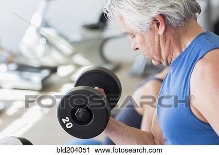 Stock Image - Older Hispanic man lifting weights in gym. Fotosearch