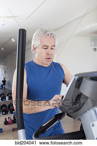 Stock Image - Older Hispanic man using elliptical machine in gym. Fotosearch