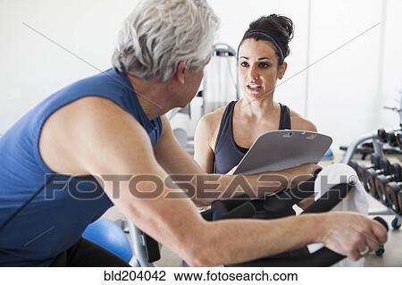 Stock Image - Older Hispanic man working with trainer in gym. Fotosearch