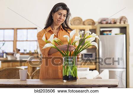 Stock Photograph - Older Hispanic woman arranging flowers in kitchen. Fotosearch