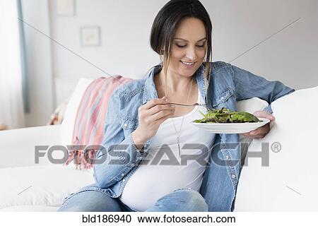 Stock Image - Pregnant Caucasian woman eating salad on sofa. Fotosearch