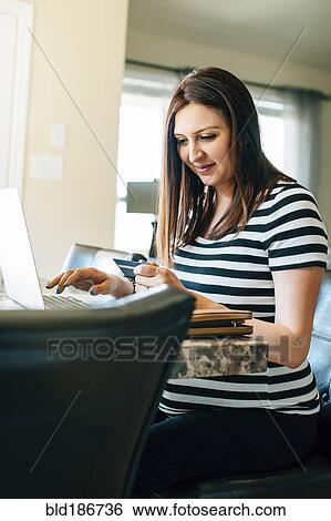 Stock Photograph - Pregnant Hispanic woman shopping with laptop in kitchen. Fotosearch