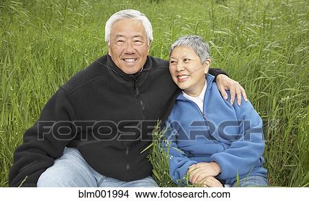 Picture - Senior Asian couple sitting in tall grass smiling. Fotosearch